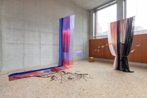 An art installation in a bright room. The installation features sheer, colourful fabrics hanging from the ceiling to the floor in a minimalist room with concrete walls and large windows.