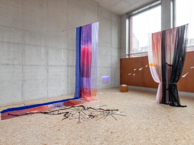 An art installation in a bright room. The installation features sheer, colourful fabrics hanging from the ceiling to the floor in a minimalist room with concrete walls and large windows.