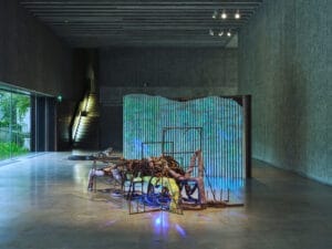 An art installation in a dark room. It consists of a metal frame structure with organic materials draped and intertwined within the frame. In the background, a textured room divider with blue, glooming vertical lines.